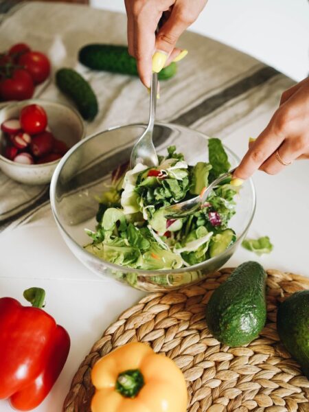 Hands preparing a fresh vegetable salad with cucumbers, peppers, and lettuce in a kitchen setting.