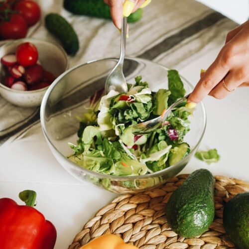 Hands preparing a fresh vegetable salad with cucumbers, peppers, and lettuce in a kitchen setting.