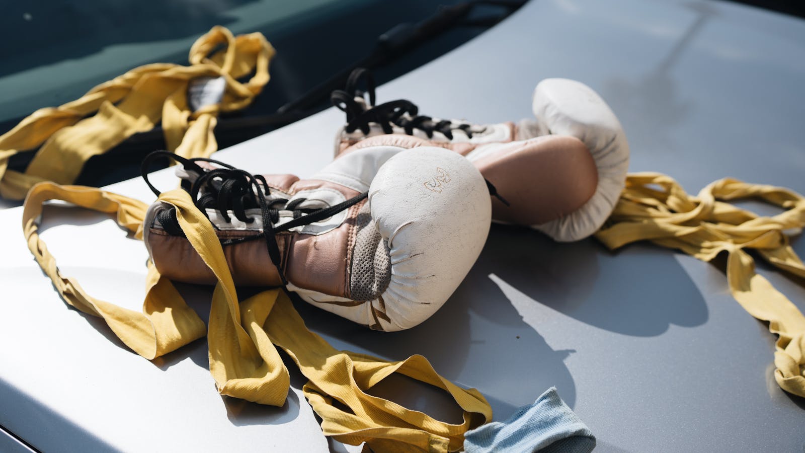 White boxing gloves and yellow wraps laid on a car hood under sunlight.